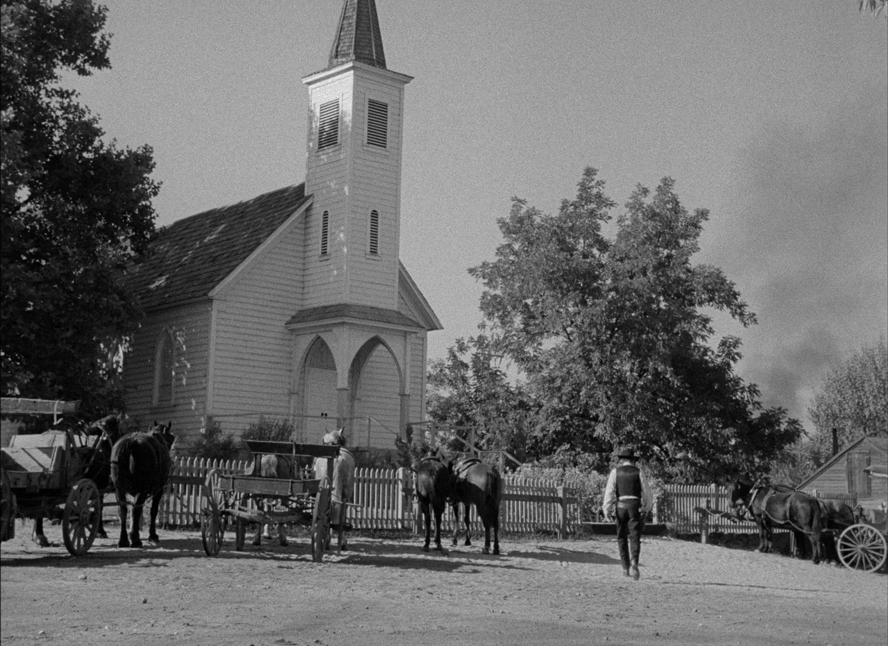 High Noon (1952) movie still: wide — A rural wooden church stands next to several horse-drawn wagons on a dirt road. A solitary man wearing a…