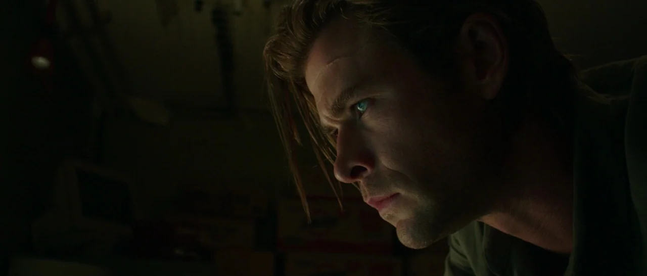 Blackhat (2015) movie still: closeup — A dimly lit, shadowed room filled with boxes in the background. A man looks toward a light source from the…