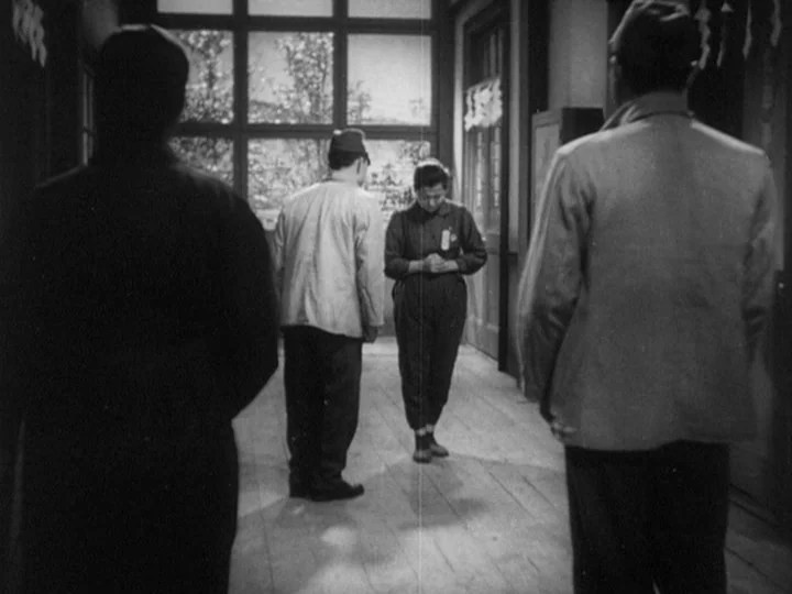 The Most Beautiful (1944) movie still: fullbody — A wooden-floored corridor features a large grid-pattern window at the far end. Three people stand…