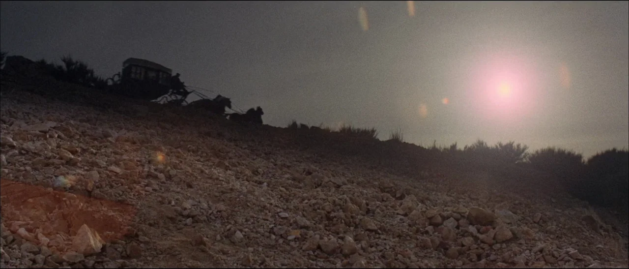 The Grand Duel (1972) movie still: wide — A rugged, desolate rocky landscape rises toward a hazy, sun-filled sky. A horse-drawn carriage moves…