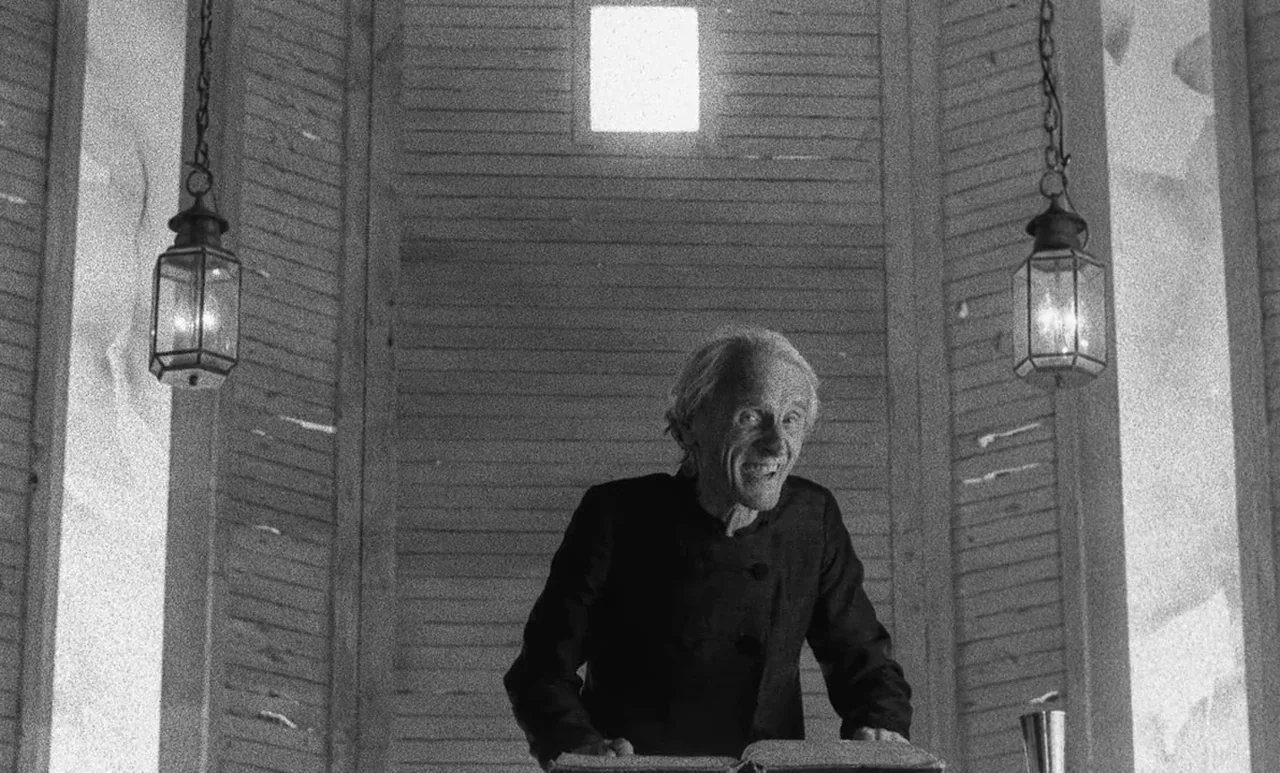 Go Down Death movie still: medium — A wooden pulpit interior features horizontal slat walls and two hanging metal lanterns. An older man leans over…