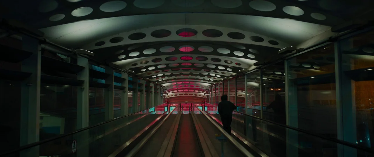Titane (2021) movie still: wide — A long, enclosed pedestrian walkway features curved ceilings with circular cutouts and glass walls. A solitary…