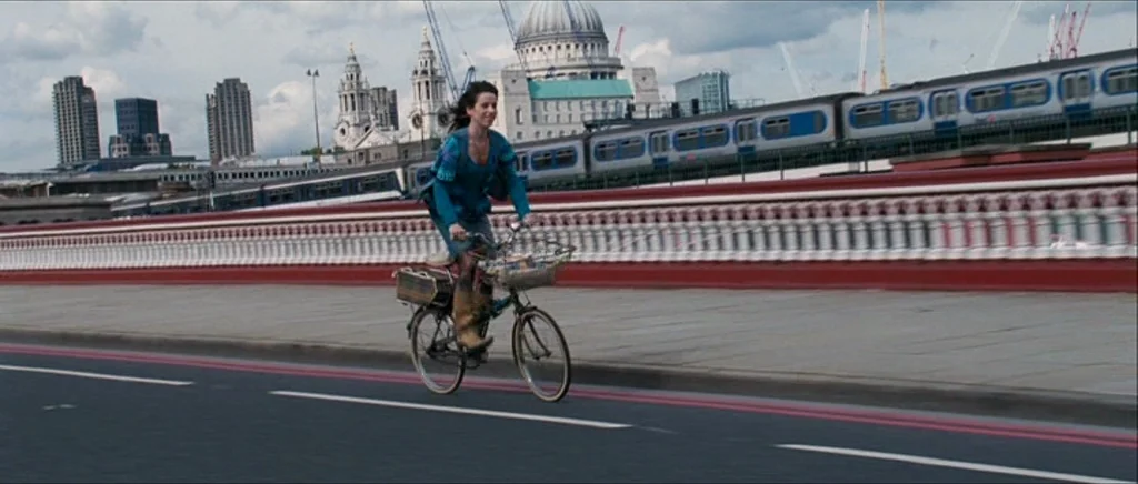 Happy-Go-Lucky (2008) movie still: fullbody — A woman rides a bicycle along a bridge over a city landscape. A large domed cathedral and a commuter…