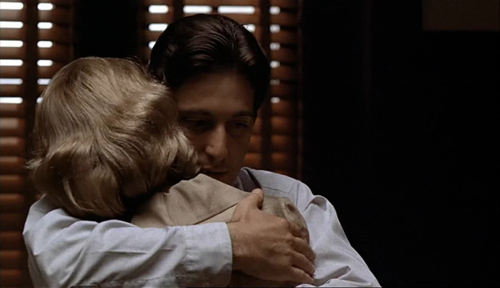 The Godfather (1972) movie still: medium — A dimly lit room features a window with dark wood slatted blinds. A man in a white dress shirt holds a…