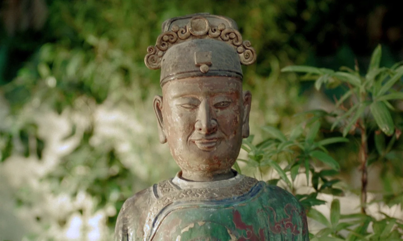 The Scent of Green Papaya (1993) movie still: closeup — A weathered wooden sculpture stands outdoors with green foliage softly blurred in the…