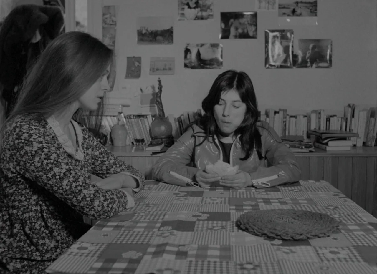 Je Tu Il Elle (1974) movie still: medium — Two women sit across from each other at a kitchen table with a checkered cloth. Photographs are pinned to…