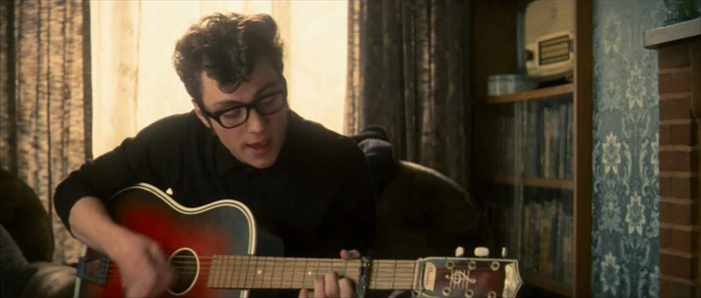 Nowhere Boy movie still: medium — A room with patterned blue wallpaper, a brick fireplace, and a wooden shelf. A young man sits while playing a red…