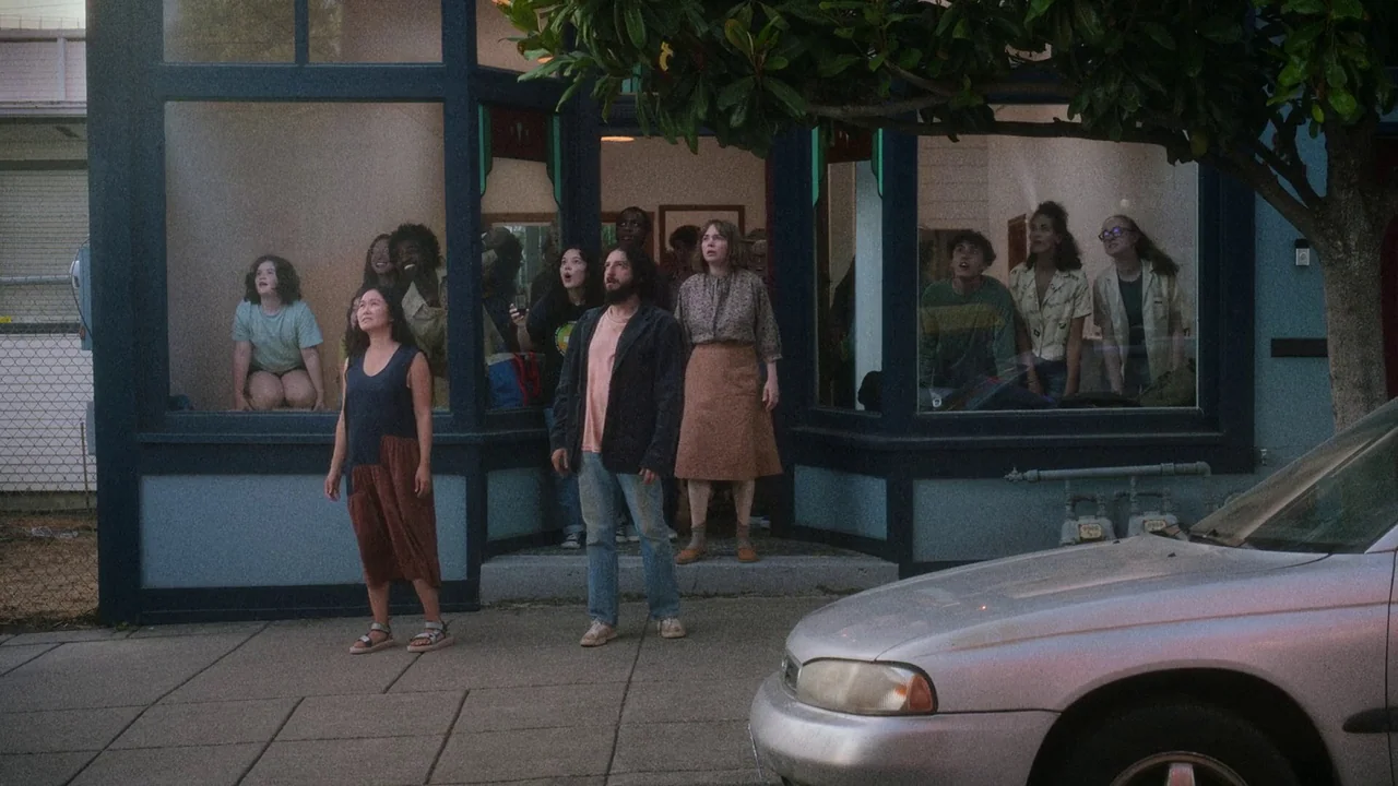 Showing Up (2022) movie still: wide — A group of diverse people stand outside a blue-trimmed storefront at dusk. They watch something in the sky…