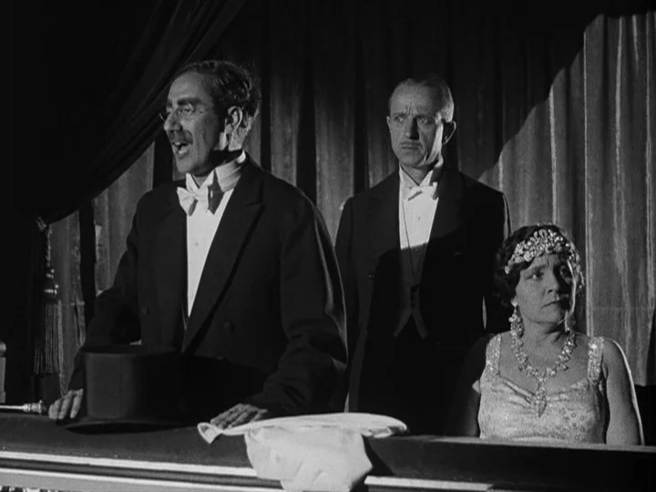 A Night at the Opera (1935) movie still: medium — A dimly lit theater box featuring three people positioned in front of heavy velvet curtains. The…
