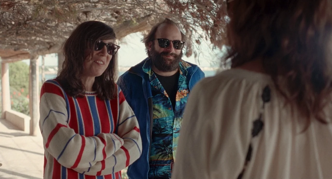 Non-Fiction (2018) movie still: medium — A man and woman stand on an outdoor patio beneath a slatted roof structure. They are turned to face someone…