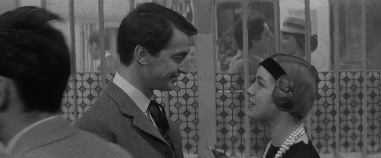 Jules and Jim (1962) movie still: medium — A cafe interior with patterned glass panels forming a decorative backdrop. A man in a suit looks toward a…