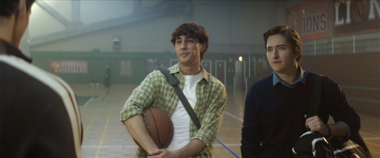 Tell Me Softly (2025) movie still: medium — A high school gymnasium interior with wooden flooring and green wall panels. Two young men stand in the…