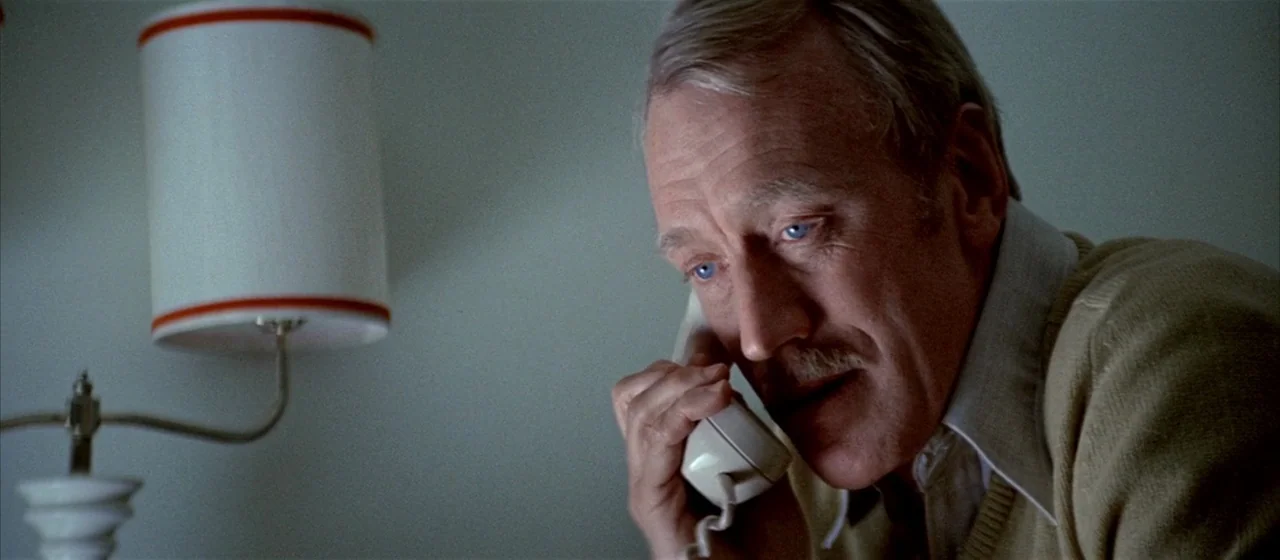 Three Days of the Condor (1975) movie still: closeup — A man speaks on a telephone next to a lamp in a dimly lit room. The wall is a plain light…