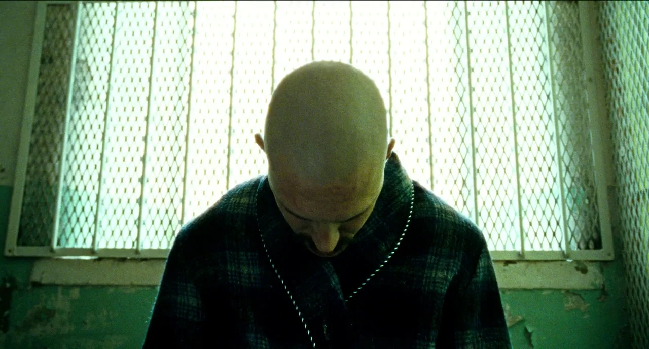 Bronson (2008) movie still: medium — A spartan space with a metallic window grate blocking daylight against the back wall. The bald man faces away…