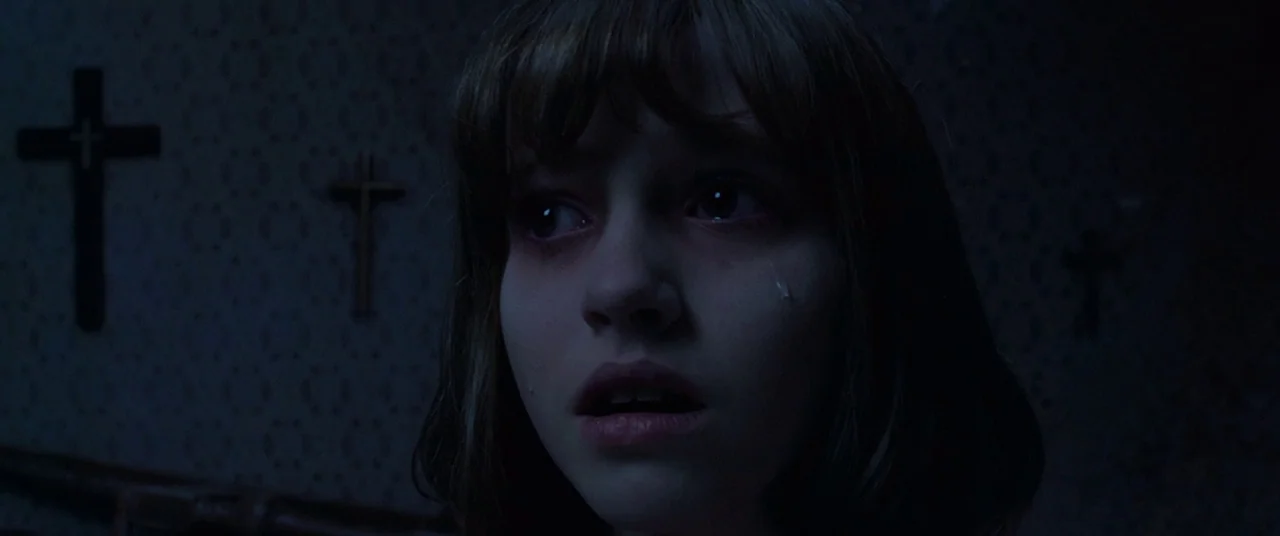 The Conjuring 2 (2016) movie still: closeup — A dimly lit room with patterned wallpaper featuring dark wooden crosses mounted on the wall. A young…