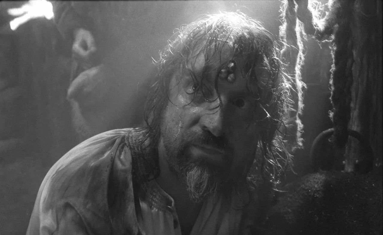 Hard To Be A God (2013) movie still: closeup — A dark, cramped interior setting with rough textures and thick ropes visible overhead. A man with…