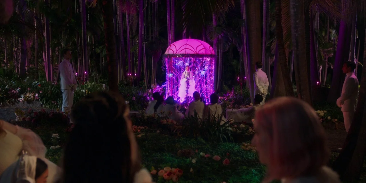 Paradise Hills (2019) movie still: wide — A tropical palm grove is lit at night by an illuminated pink and purple structure. A person stands inside…