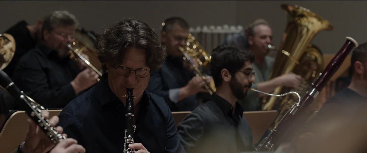 Tár (2022) movie still: medium — A group of musicians sits together in a rehearsal space with muted wood surfaces. The central clarinetist plays his…