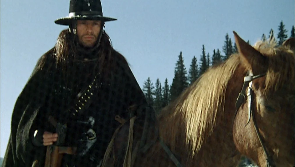 The Great Silence (1968) movie still: medium — A man on horseback moves through a cold mountain pass with coniferous trees. The man wears a dark…