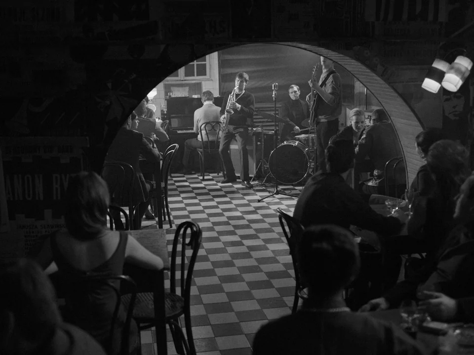 Ida (2013) movie still: wide — A dimly lit club features an arched doorway looking toward a central performance stage. A four-piece band plays…