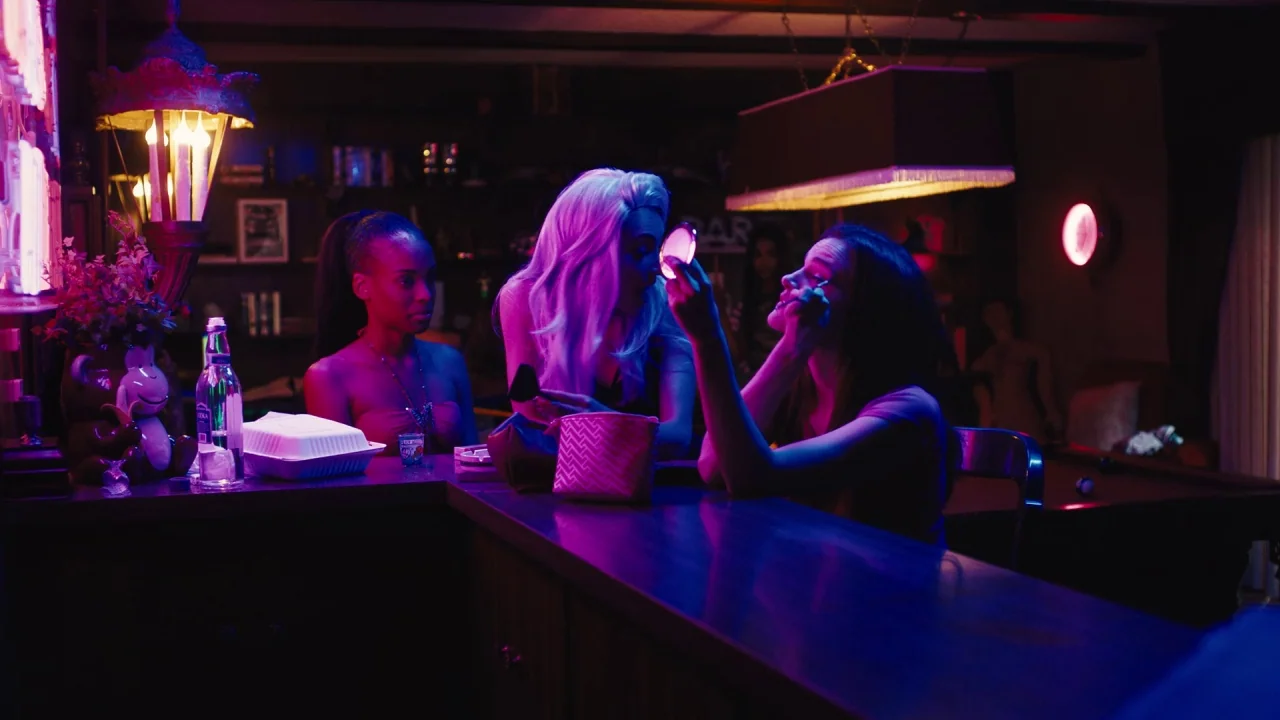 Cam (2018) movie still: medium — A dark bar interior features a wooden counter and shelves lined with bottles. Three young women sit together in…