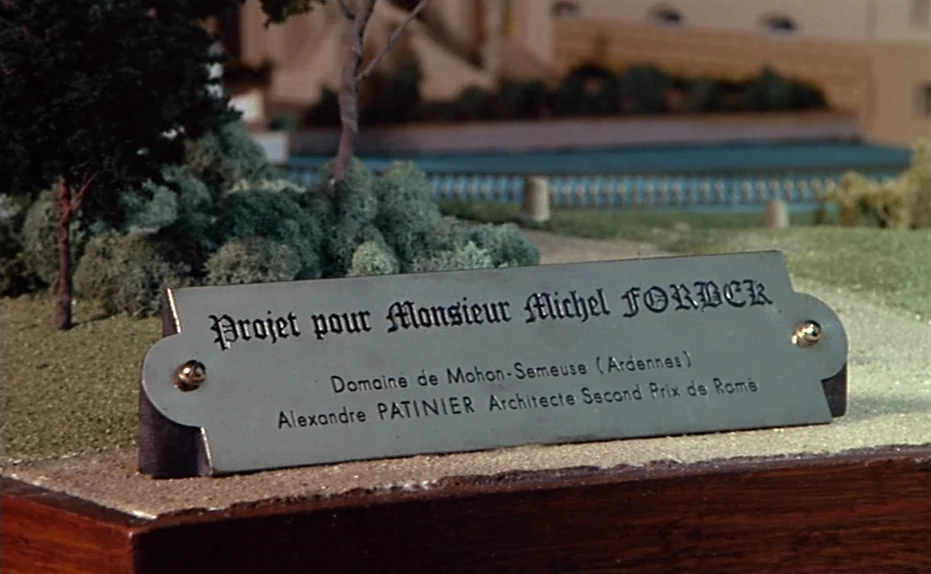 Life Is A Bed of Roses (1983) movie still: closeup — A close-up of a metal plaque on a miniature architectural model base. The plaque identifies the…