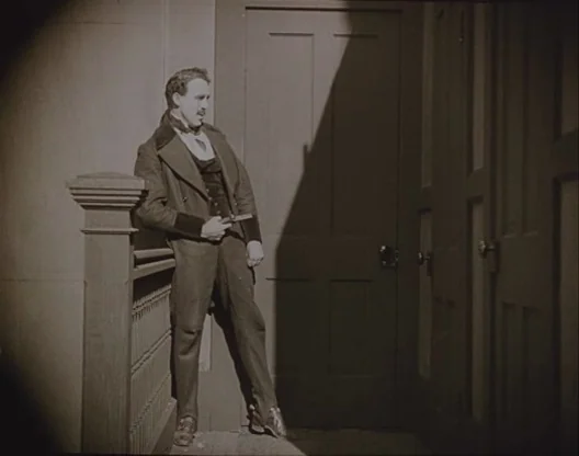 Birth of a Nation (1915) movie still: fullbody — A stairwell landing features a tall wooden door and a decorative banister. The man rests against…