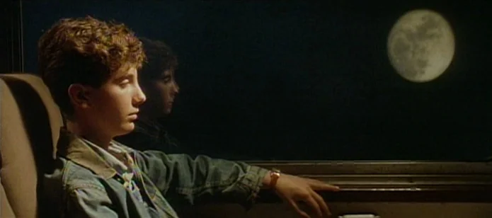 The Neon Bible (1995) movie still: medium — A boy sits inside a darkened vehicle during the night hours. He gazes through a glass window toward a…