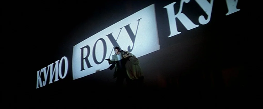 Anguish (1987) movie still: wide — Two individuals stand against a large, brightly lit sign reading ROXY at night. The man points a gun while the…
