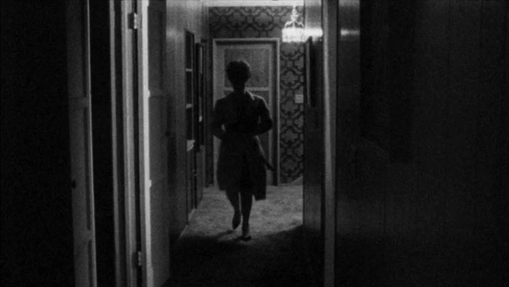 Faces (1968) movie still: fullbody — A residential hallway with dark walls and patterned wallpaper leads to a distant doorway. A woman walks toward…