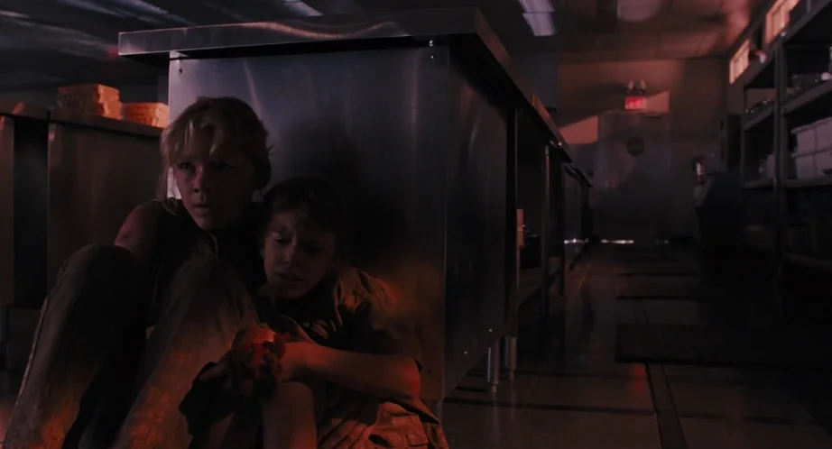 Jurassic Park (1993) movie still: medium — A dark industrial kitchen interior features large stainless steel counters and metal shelving. Two young…