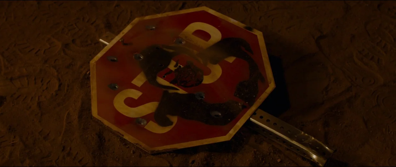 El Chicano (2018) movie still: closeup — A damaged metal stop sign rests on a dusty, uneven desert floor. The sign has multiple bullet holes and…