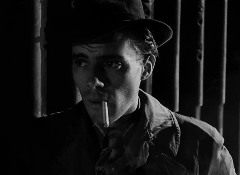 The Blue Lamp (1950) movie still: closeup — A man stands in a dark space with vertical iron bars in the background. He holds a cigarette in his…