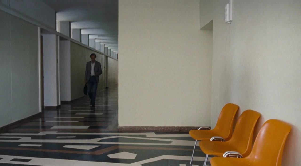 Paradise: Hope (2013) movie still: wide — A long, sparsely furnished corridor features patterned linoleum flooring and pale walls. A man walks…