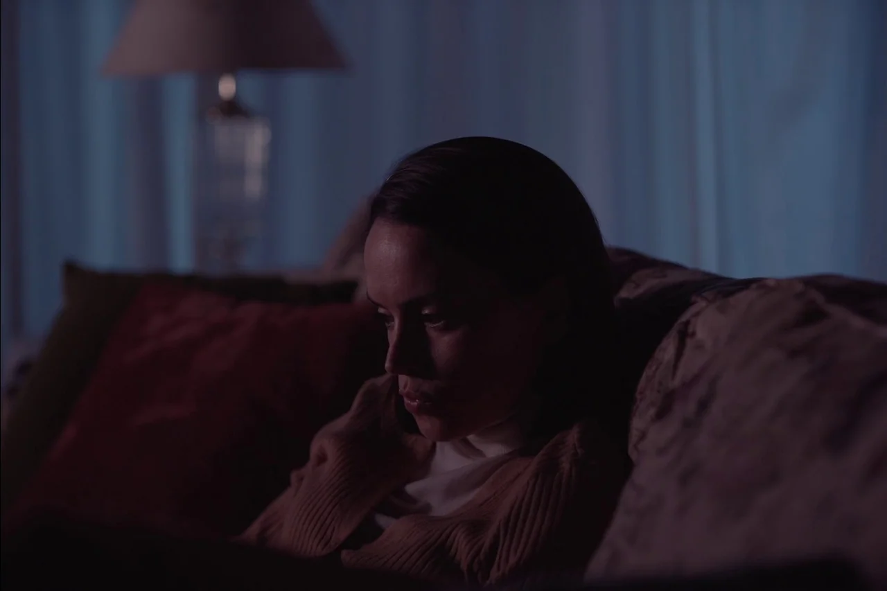 Sometimes I Think About Dying (2023) movie still: medium — A dark living room features a person seated on a sofa at night. The subject is looking…