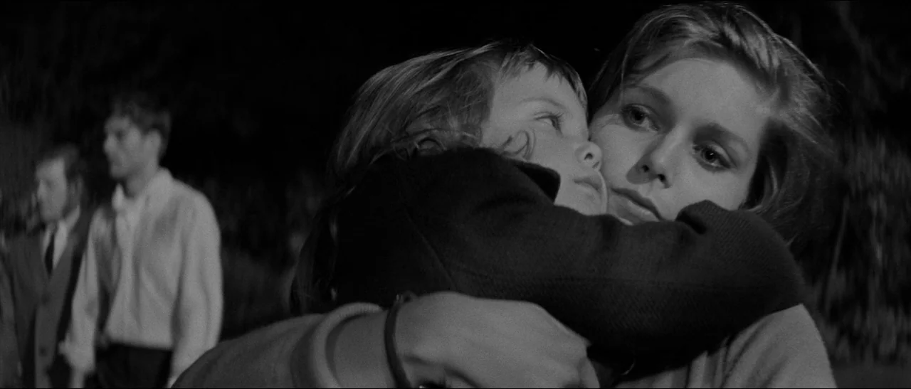 Bunny Lake Is Missing (1965) movie still: closeup — An outdoor nighttime setting features a woman embracing a small child in close focus. Blurred…