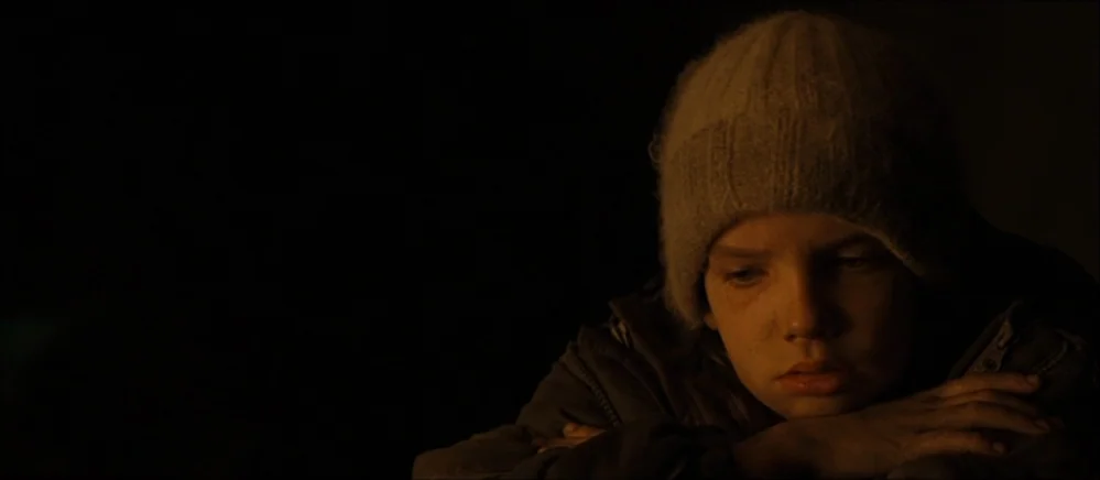 The Road (2009) movie still: closeup — A dark, dimly lit environment where only the subject is visible. A young child wearing a knit hat rests their…