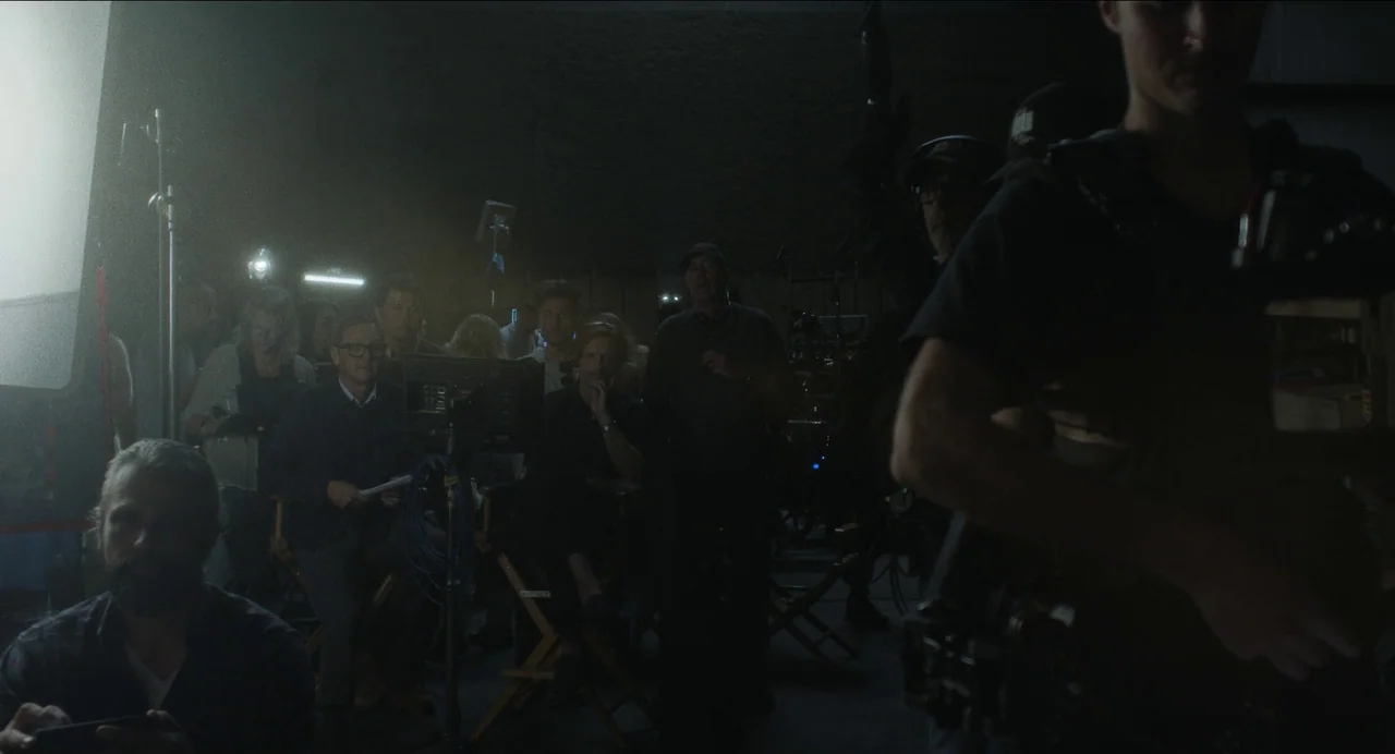 May December (2023) movie still: medium — A dimly lit production set interior with a group of crew members sitting down. Several people are watching…