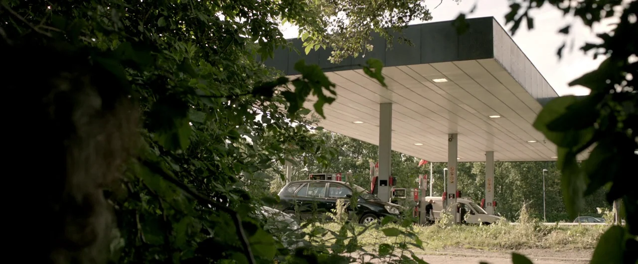 Borgman (2013) movie still: wide — A modern roadside petrol station sits surrounded by dense green foliage and deciduous trees. Several parked cars…