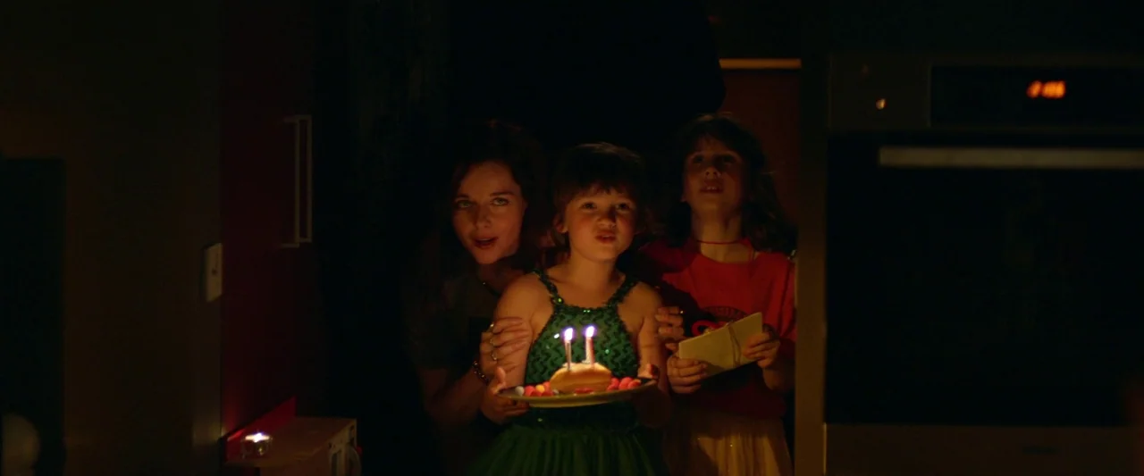 Sibyl (2019) movie still: medium — A dark indoor space illuminated by the glow of small candles on a cake. Three individuals stand together in a…