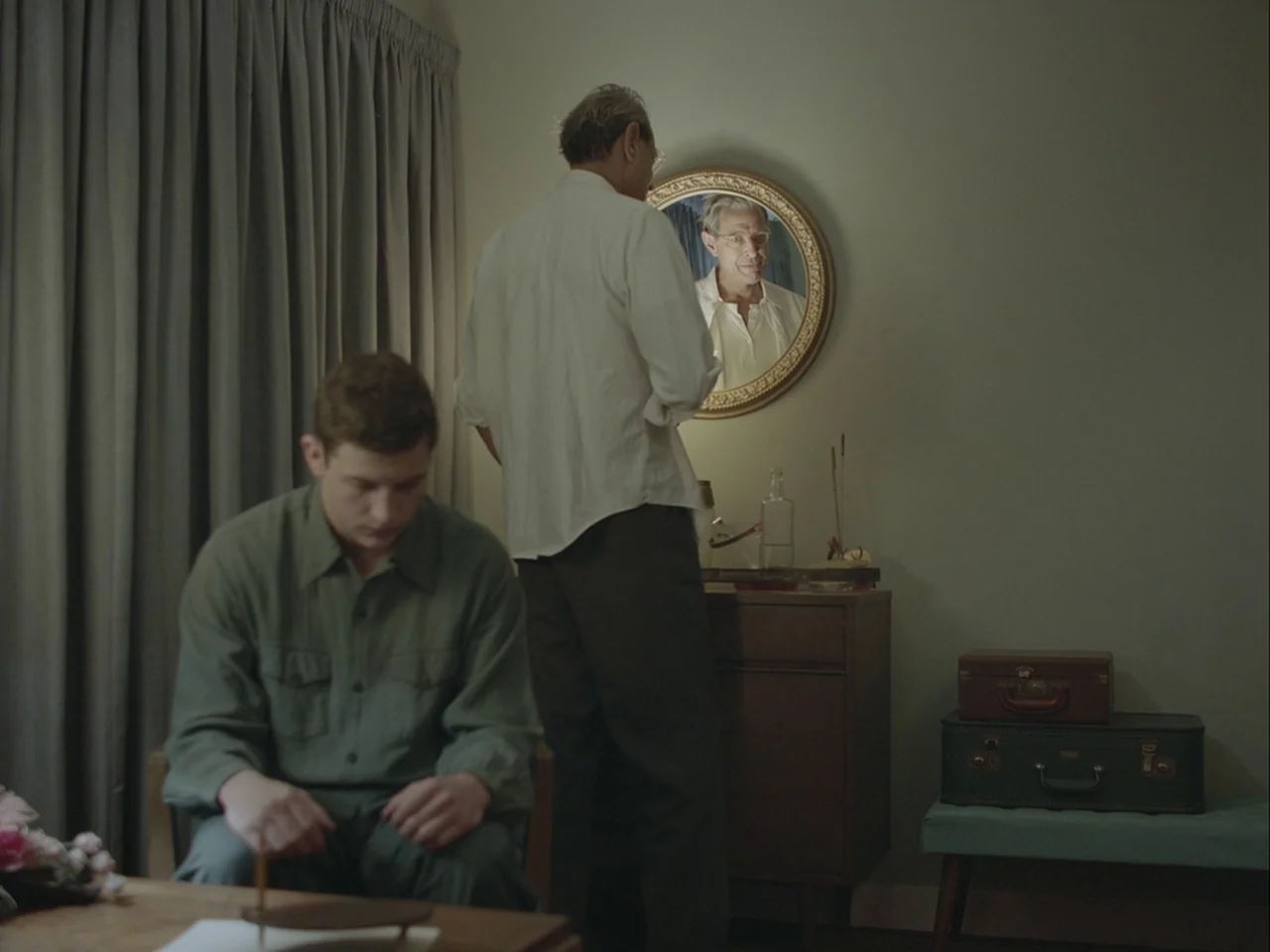 The Mountain (2018) movie still: medium — A spare, mid-century motel room features drab green walls, grey curtains, and vintage luggage. An older…