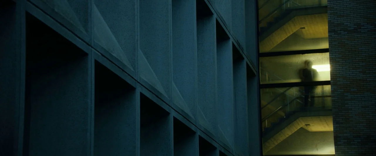 The Report (2019) movie still: wide — A brutalist concrete exterior features a glass-enclosed stairwell illuminated from within by artificial light.…