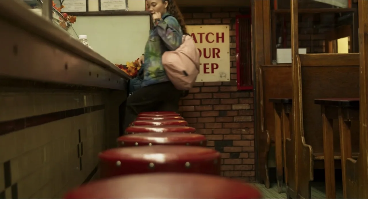 Salvation (2025) movie still: medium — A diner interior features a row of red vinyl stools along a long counter. A young woman with a pink backpack…