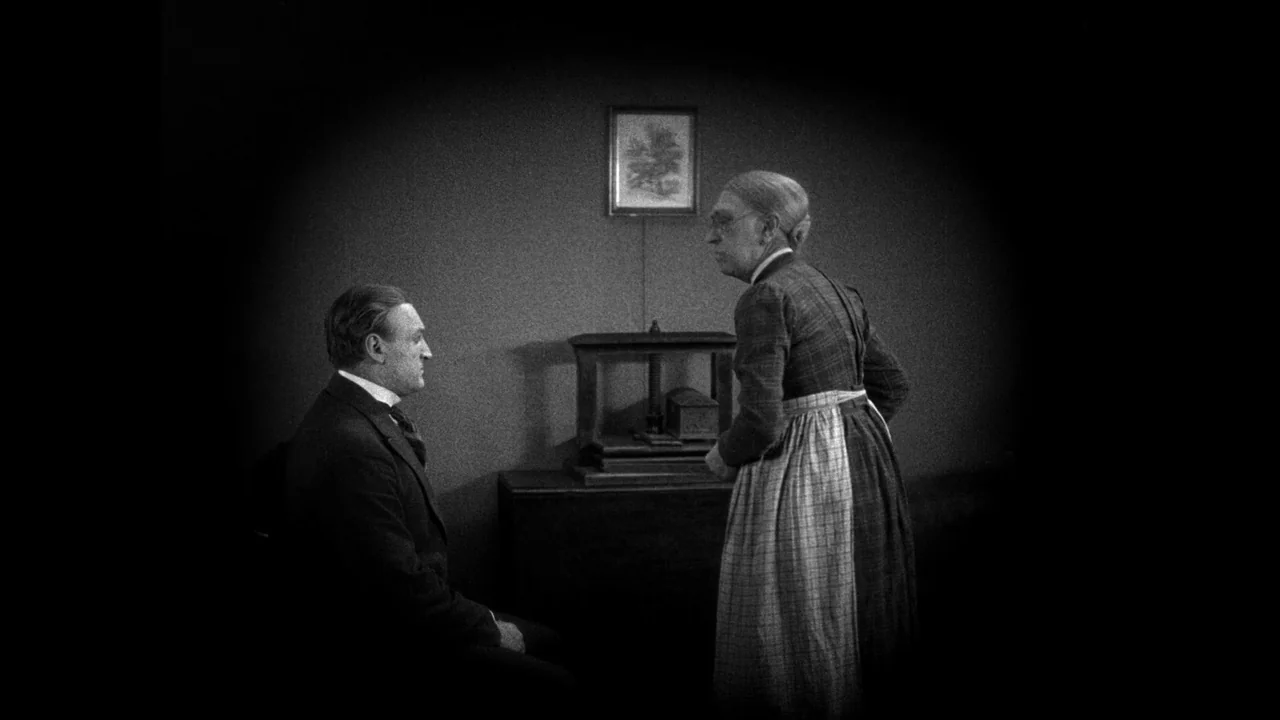 Master of the House (1925) movie still: medium — A sparsely furnished 1900s room features a wooden side table and wall-mounted picture frame. An…