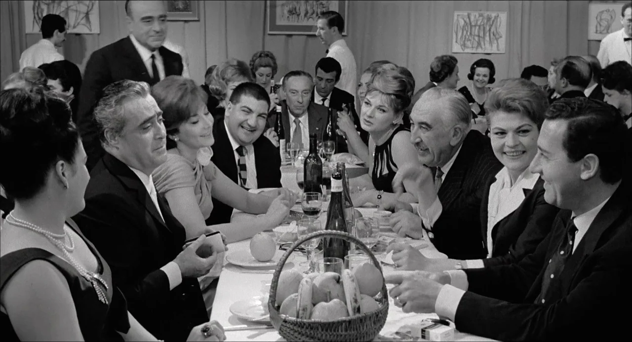 Il Boom (1963) movie still: medium — A crowded restaurant interior with guests seated around a long banquet table. Men and women in formal attire…