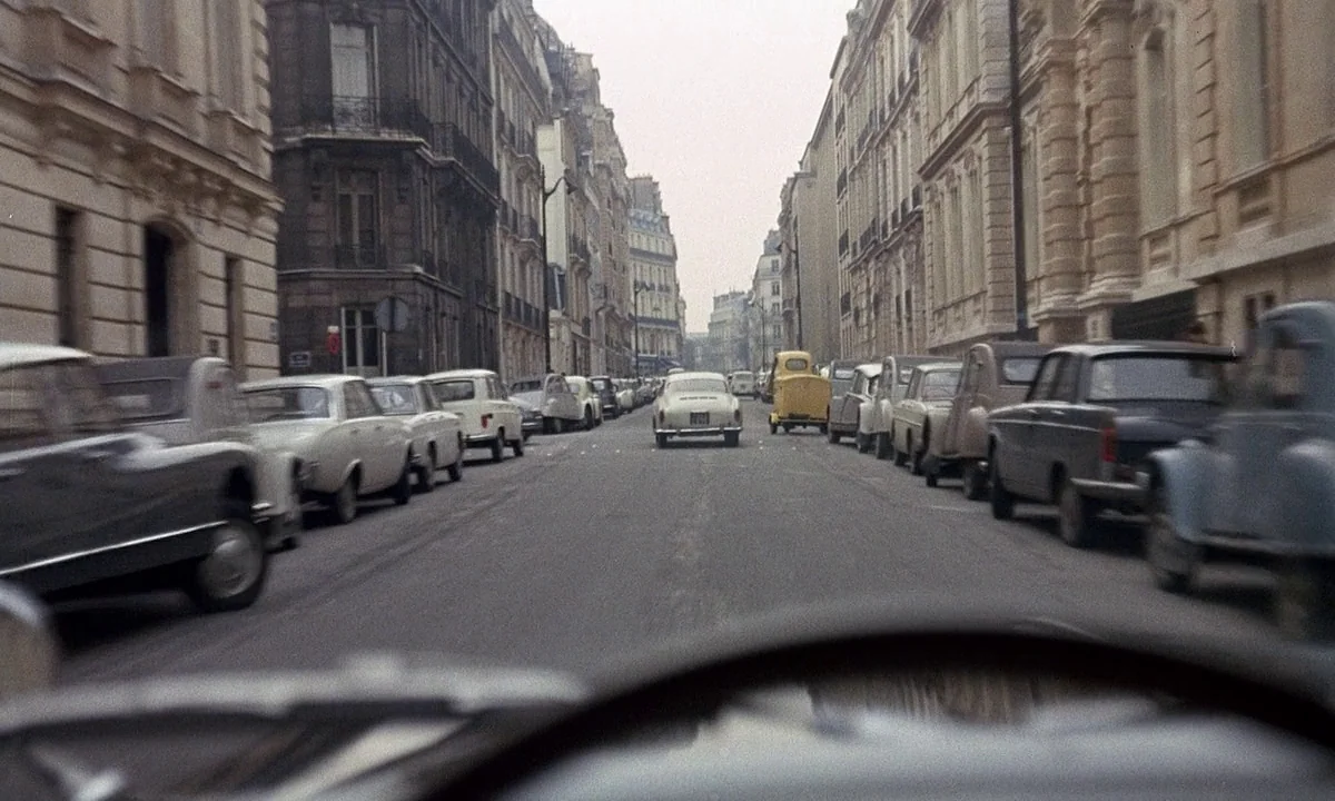 Belle De Jour (1967) movie still: establishing — A narrow Parisian-style street is lined with multi-story stone buildings and tightly parked vintage…