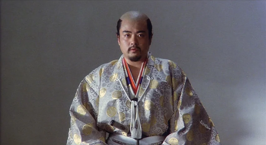 Kagemusha (1980) movie still: medium — A man stands centered against a smooth, solid-colored wall inside a minimalist room. He wears traditional…