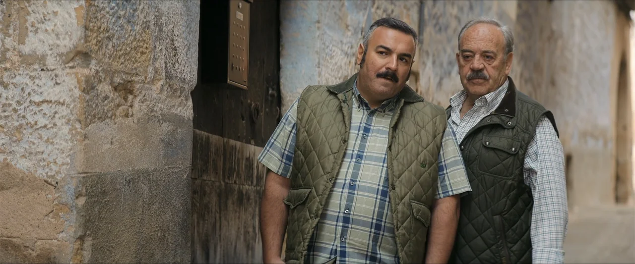 A Hipster in Rural Spain (2024) movie still: medium — A narrow alley with textured, weathered stone walls and an old wooden door. Two middle-aged…