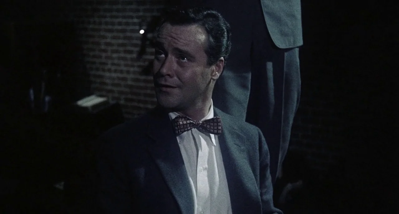 Bell Book and Candle (1958) movie still: medium — A dimly lit brick-walled space with dark, shadowy corners. A man in a suit looks toward an unseen…