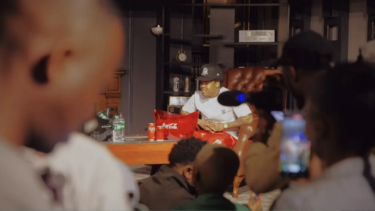 African Throne World Tour (2024) movie still: medium — A studio setting with a person seated in a brown leather chair. Various drinks are arranged…